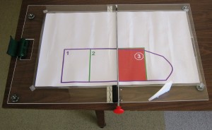 Figure 1: The pillowcase folding aid, which includes the acrylic work surface, a center divide rode that provides a central axis for folding. One of the folding templates is shown on the work surface, which outlines the different steps for folding a particular pillowcase.