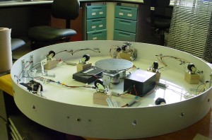 FFigure 2.  Lazy Susan with the top disc removed.  The DC motor is in the center, along with the electronic circuitry box.  There are 10 casters distributed around the device to hold the weight of the top disc.  LEDs are along the perimeter.igure 2. Lazy Susan with the top disc removed. The DC motor is in the center, along with the electronic circuitry box. There are 10 casters distributed around the device to hold the weight of the top disc. LEDs are along the perimiter.