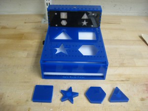 Figure 1: The Boxxy device with four of the shapes.  When the device detects a shape going down the slide, it illuminates the appropriate icon on the back panel.  The shape then slides out of the slot on the front edge.