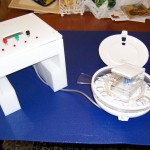 Figure 1: Front View of EMOD. The Pill Dispenser is on the right with its lid open. The Pill Loader and Pill Cutter are on top of the dispenser, and the mounting stand is on the left.